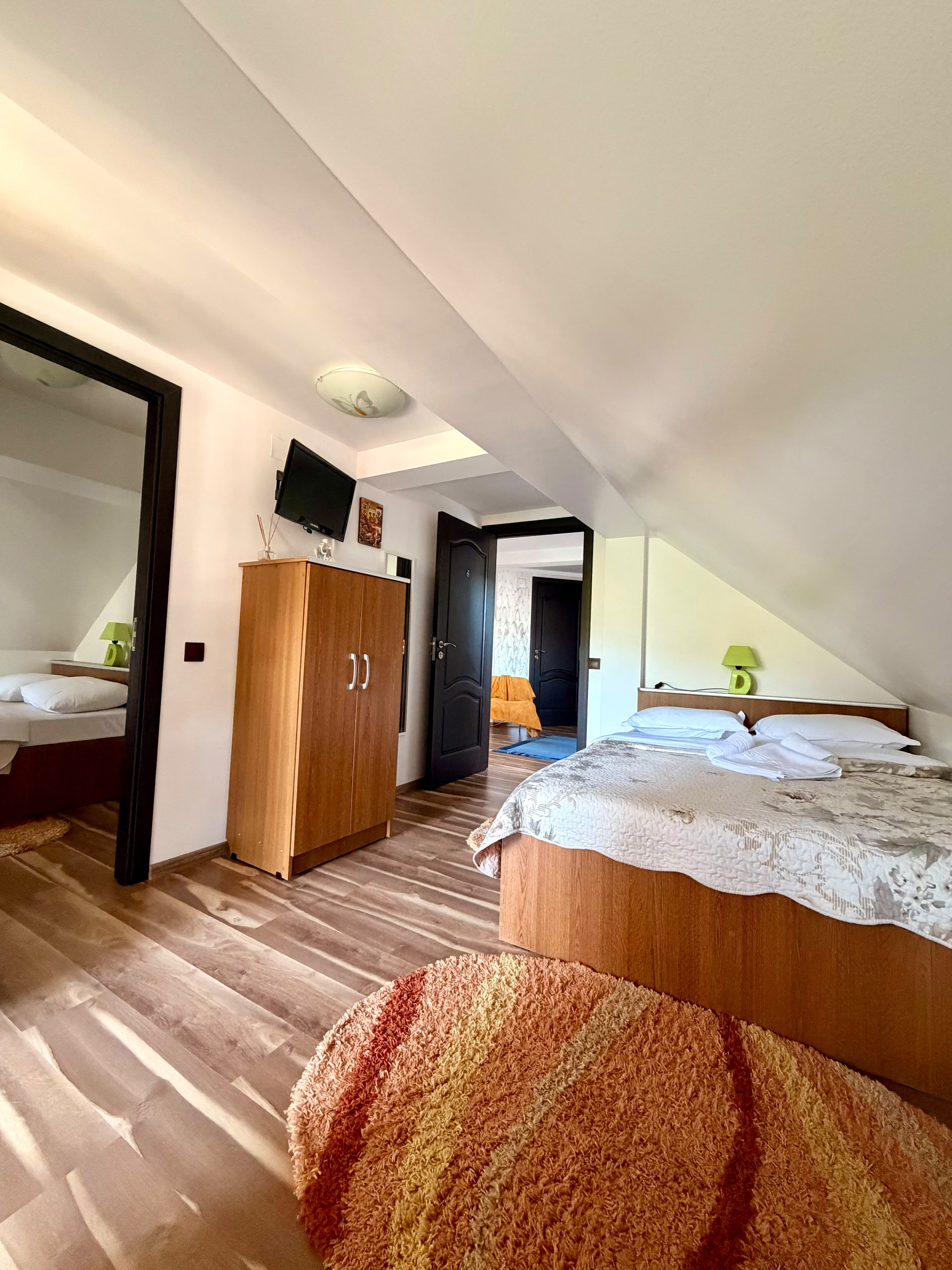 Prima cameră a apartamentului de la mansardă, cu pat matrimonial, televizor și ușa deschisă spre a doua cameră