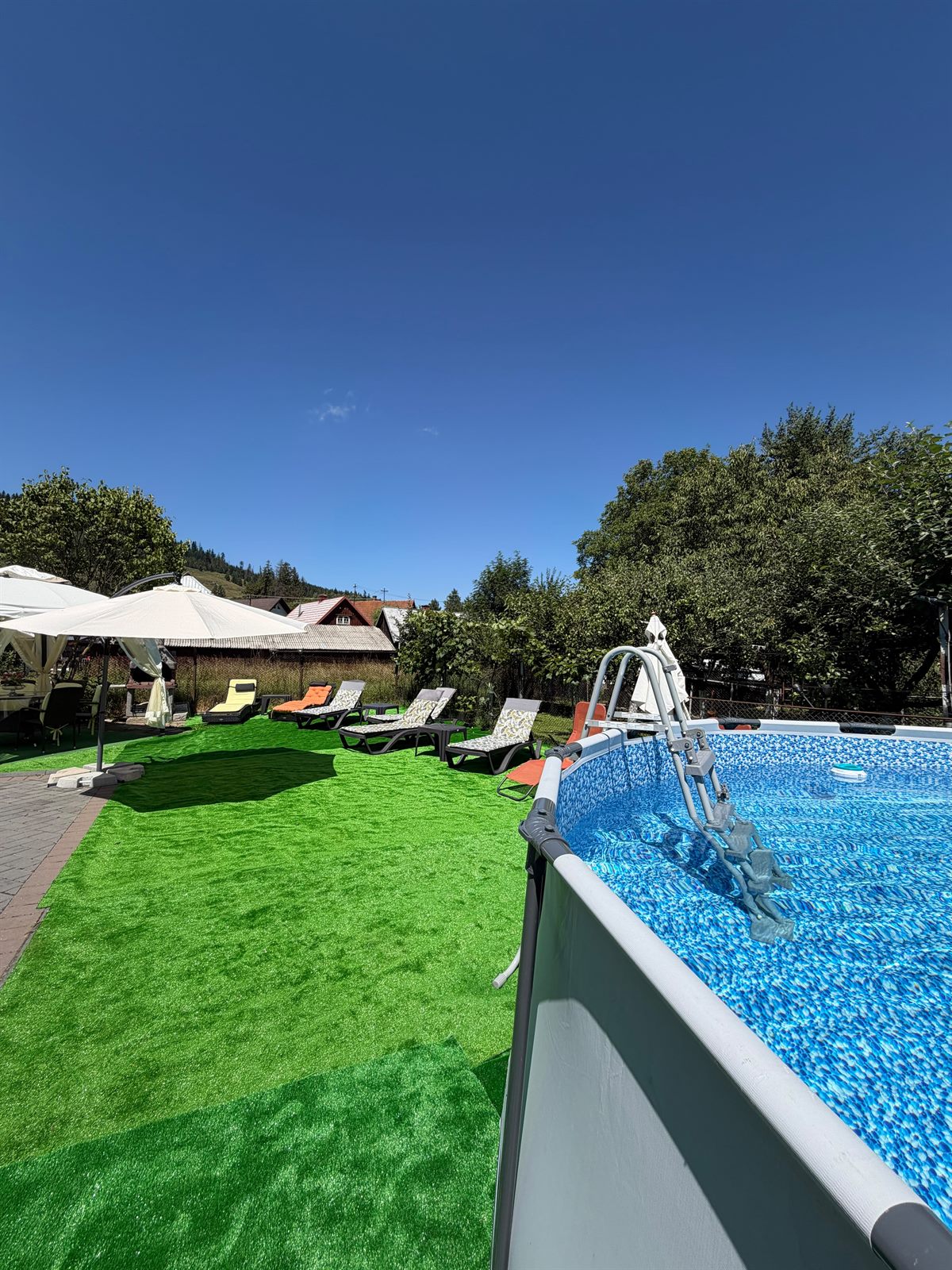 Zona de piscină văzută dinspre casă, cu gazon, șezlonguri și umbrelă mare